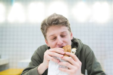 Genç bir adam hafif bir restoranda lezzetli bir hamburger yiyor. Aç öğrenci zevk ile hamburger yiyor