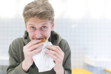 Komik de genç bir öğrenci bir sandviç ısırıkları ve kameraya benziyor. Bir hamburger yeme genç bir adam portresi.
