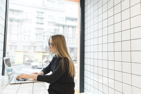 La chica rubia se sienta en un elegante restaurante cerca de la ventana y trabaja para un ordenador portátil. Estudiantes estudian en una cafetería cerca de la ventana — Foto de Stock