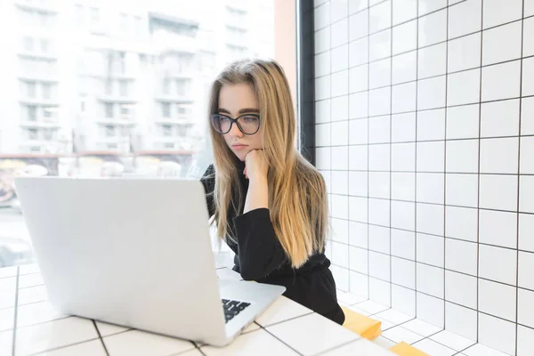 Uma bela estudante goza de um laptop em um café elegante, moderno e leve. A menina se senta no restaurante na janela e usa a internet . — Fotografia de Stock