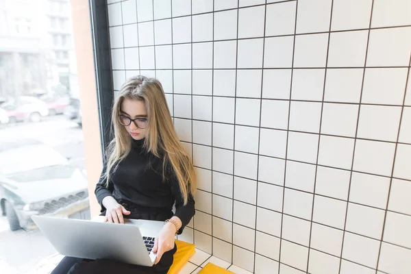 Fiatal lány ül a laptop a kávézóban kör és a munka. Egy diák lány használja az internetet a laptop a kávézóban az ablak mellett — Stock Fotó