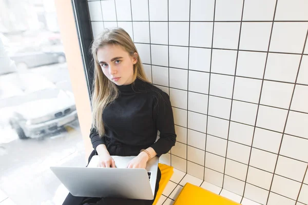 Портрет молодої дівчини, що сидить з ноутбуком на колінах і кафе біля вікна. Подивіться на камеру. Студентські заняття в яскравому стильному кафе — стокове фото