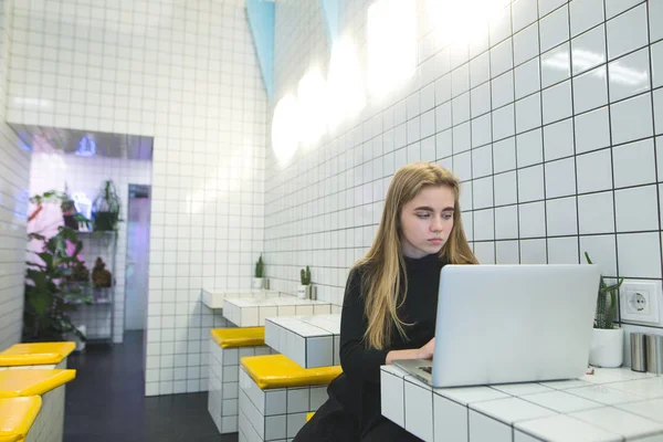 La ragazza usa internet su un computer portatile in un caffè moderno. Gli hipster lavorano su un computer portatile in un caffè con un interno elegante. Lavoro su un computer portatile in un luogo pubblico — Foto Stock