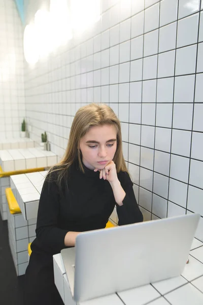 Una giovane ragazza utilizza internet su un computer portatile in un caffè con un interno elegante. Lavoro su Internet in un caffè moderno . — Foto Stock
