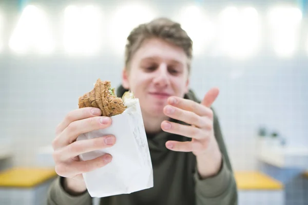 A kezében egy közeli Sedvich. Egy pozitív ember eszik egy hamburgert egy elegáns, könnyű, gyorsétterem — Stock Fotó