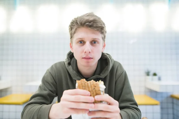 Retrato de una joven estudiante guapa con una hamburguesa en sus manos en un establecimiento de comida rápida elegante y brillante. Un joven con una hamburguesa mira a la cámara en sus manos — Foto de Stock
