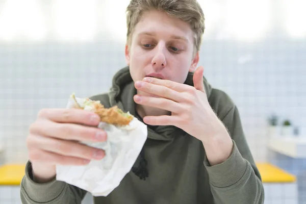 El estudiante se lame los dedos y mira hambriento mirando la silla de montar en sus manos. Joven come una hamburguesa en un restaurante de comida rápida . — Foto de Stock