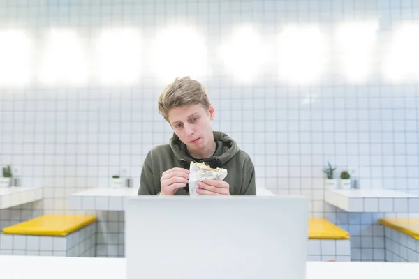 Un joven come un sándwich y usa un portátil en una cafetería. Trabajar durante un descanso para almorzar en un restaurante de comida rápida —  Fotos de Stock