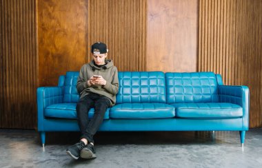 Mavi bir kanepede oturan ve bir smartphone kullanarak genç adam. Öğrenci kanepede oturan ve üzerinde esmerleri bir ileti yazar. Ahşap duvar arka plan üzerinde.