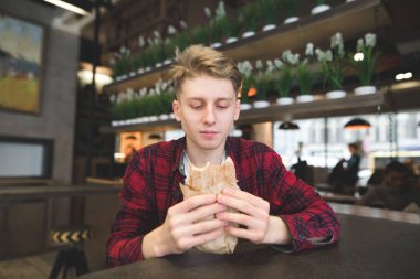 Aç bir göz ile bir öğrenci onun elinde sandviç bakar. Genç adam ile bir kafede hamburger yemek.