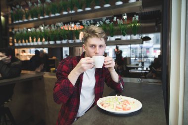 Genç bir adam kahve içer ve rahat bir ördek bir salata yiyor. Bir fincan kahve ve salata ile öğrenci yemekleri. Bir fincan kahve ve salata ile öğrenci yemekleri. Kameraya bak