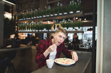 Kırmızı gömlekli genç bir öğrenci bir salata ve rahat bir kafede kahve ile yiyor. Güzel bir adam o çatal salata yiyor. Kafede Öğle Yemeği