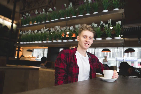 El joven positivo se sienta en un café con una taza de café y sonríe. Café en la cafetería — Foto de Stock