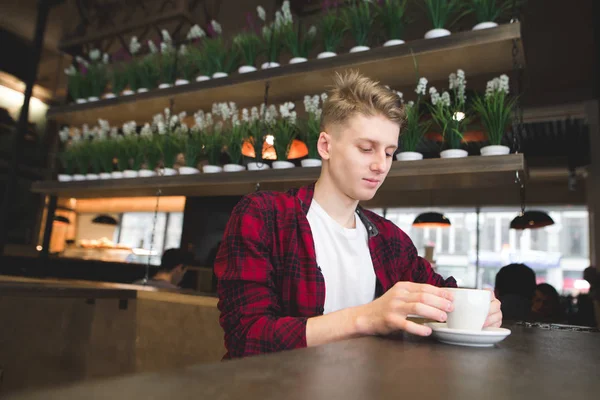 Stylish young man in a red trash sits in a cafe with a cup of coffee. Breakfast A look at a drink at a cafe. — Stock Photo, Image
