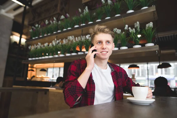 Un estudiante habla por teléfono en el café y sonríe. Un joven bebe café y habla por teléfono. — Foto de Stock