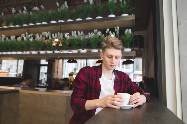 Задумчивый молодой человек сидит в кафе с пончиком кофе. Посмотрите на напиток в уютном кафе — стоковое фото