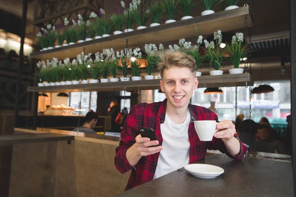 Un joven positivo sostiene una taza de café en sus manos y mira a la cámara. Un estudiante bebe café y usa un teléfono inteligente en un acogedor café — Foto de Stock