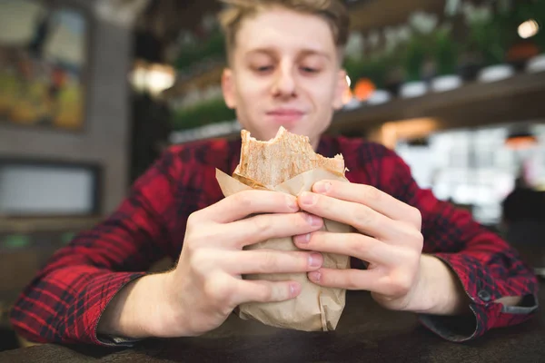 Um estudante come uma sandes num café aconchegante. Um jovem com um olhar feliz olha para a sanduíche panini. nas mãos deles. Concentra-te no sanduíche — Fotografia de Stock