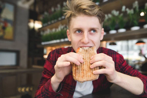 Um jovem lindo come uma sanduíche de panini em um café acolhedor. Estudante engraçado morde sanduíche. Almoço no café . — Fotografia de Stock