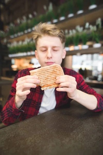 Um jovem senta-se num café e segura uma sanduíche de panini. Um estudante lava um sanduíche em um café . — Fotografia de Stock