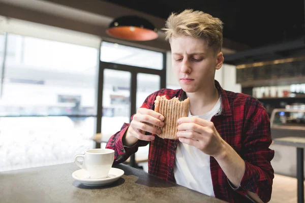 Ein gutaussehender junger Mann mit Appetit blickt beim Mittagessen in einem gemütlichen Café auf ein Sandwich in seinen Händen. Student kocht Panini-Sandwich im Restaurant — Stockfoto