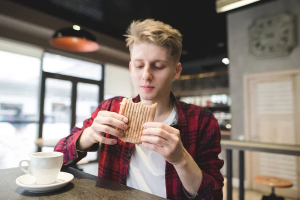 Giovane pranzo panini sandwich nel ristorante. Uno studente con appetito guardando un panino tra le mani mentre pranza in un accogliente caffè . — Foto Stock
