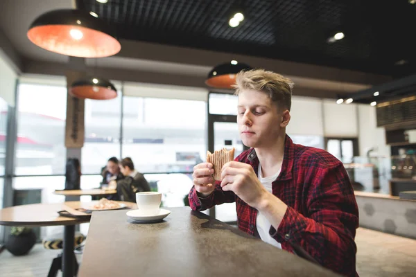 Studentengerichte mit Sandwich und Kaffee in einem gemütlichen Café. Hungriger junger Mann blickt auf den Sattel in seinen Armen. — Stockfoto