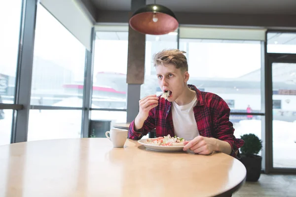 Un joven con camisa roja come una ensalada en un café cerca de la ventana. Un estudiante come corsina en un café — Foto de Stock