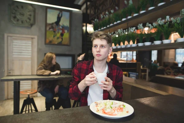 Un estudiante con una taza de café en las manos cena en un acogedor café. El joven termina mirando a un lado mientras come. — Foto de Stock