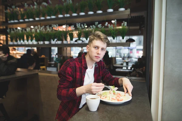 Um jovem estudante de camisa vermelha come uma salada em um café acolhedor e olha para a câmera. Um estudante come em um café — Fotografia de Stock