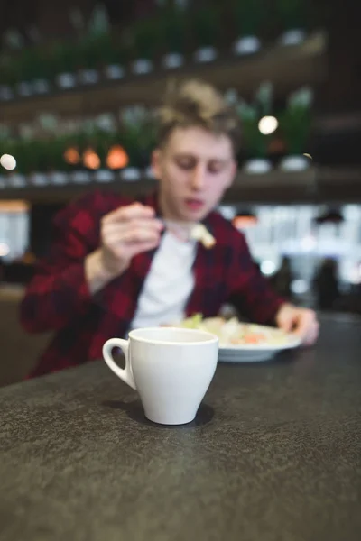 Una taza de café en el fondo de una persona que come un restaurante. La taza está enfocada, la persona está borrosa . — Foto de Stock