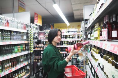 Güzel bir kız şarap şişesi dikkatle süpermarkette alışveriş yaparken görünüyor.