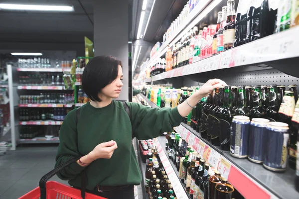 Mladá dívka vybere pivo v supermarketu. Krásná dívka nakupuje alkohol v úložišti. Nakupování v supermarketu. — Stock fotografie
