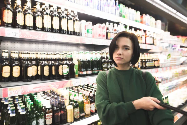 Beautiful young girl shopper posing against the background of shelves with alcohol. — Stock Photo, Image