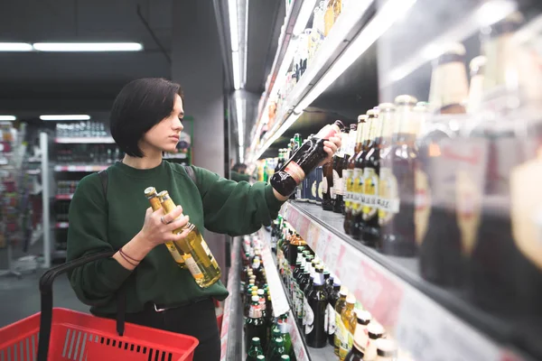 Una hermosa chica toma bebidas alcohólicas del estante del supermercado. Compras de alcohol en la tienda . — Foto de Stock