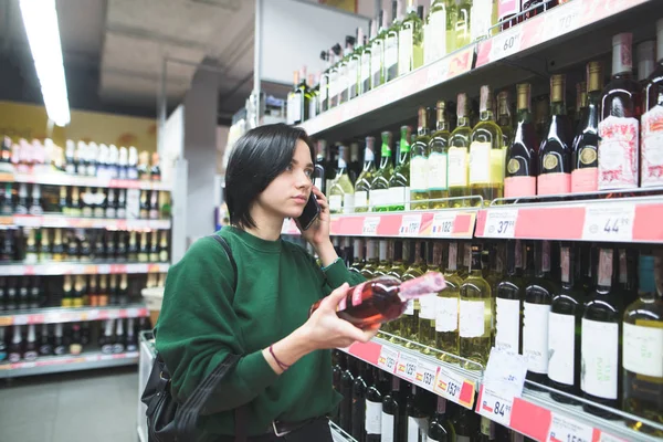 En pige med en flaske vin i hænderne taler over telefonen. En pige med en telefon køb i et supermarked . - Stock-foto