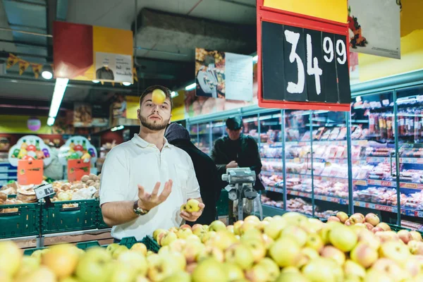 Un hombre con barba hace malabares con manzanas en el departamento de verduras del supermercado . — Foto de Stock