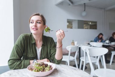 Sevimli genç kız rahat ışık restoranda salata yemek ve uzağa bakarak oturur. Kız yemekleri restoran salata ile