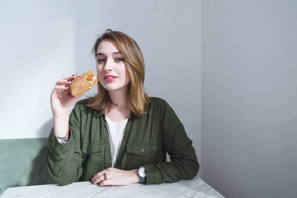 Retrato de uma menina bonita na mesa e com um sanduíche nas mãos. Uma menina com um beturbist em suas mãos olha para a câmera — Fotografia de Stock