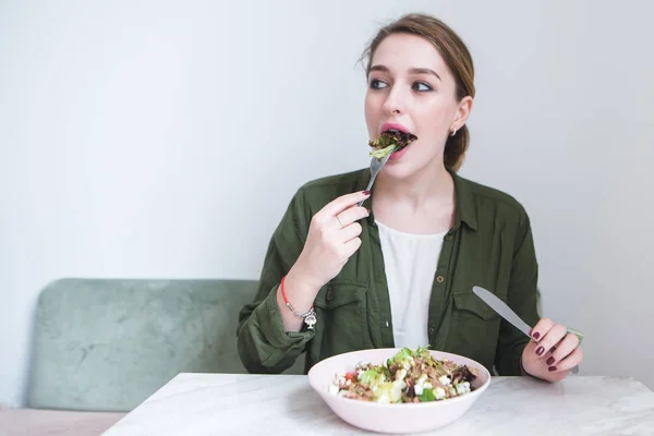 Una hermosa chica se come una ensalada de platos en un restaurante ligero y mira a un lado. La mujer muerde ensalada verde. Ensalada para el desayuno — Foto de Stock