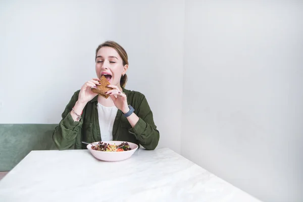 Μια όμορφη γυναίκα τρώει ένα σάντουιτς σε ένα καφενείο στο φόντο των ένα φως τοίχων. Η σκύλα δαγκώνει το σάντουιτς. Μεσημεριανό γεύμα στο εστιατόριο. — Φωτογραφία Αρχείου
