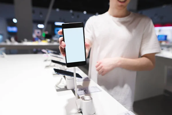 Un hombre muestra un teléfono inteligente con una pantalla blanca en la cámara. Teléfono con pantalla blanca en el fondo de la tienda electrónica. Comprar un teléfono en la tienda . — Foto de Stock