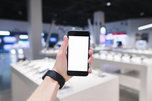 Ein Uhrzeiger hält ein Telefon mit weißem Bildschirm vor dem Hintergrund eines Elektronikgeschäfts. Kauf eines Smartphones im Geschäft — Stockfoto
