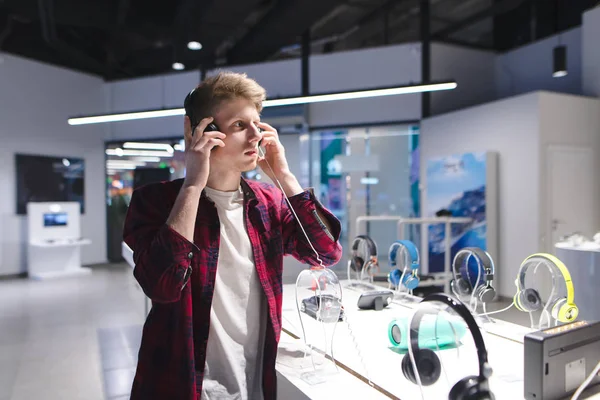 Un beau jeune homme testant des écouteurs dans le magasin d'électronique. Choisir et acheter des écouteurs dans un magasin de technologie . — Photo