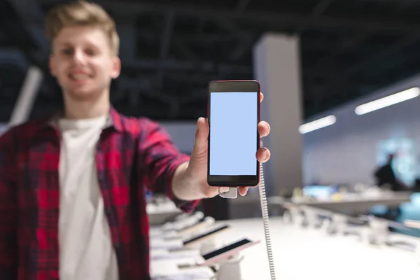 Ein positiver junger Mann schickt ein Smartphone mit weißem Bildschirm in die Kamera und lächelt. Junger Mann mit einem Handy in der Hand eines Elektronikgeschäfts — Stockfoto