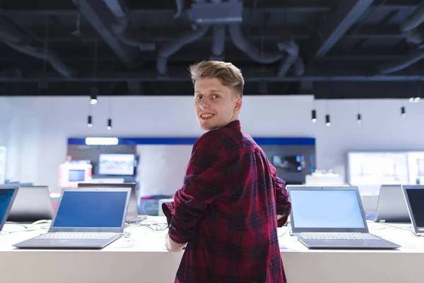 Un joven positivo en el departamento de informática de la tienda de electrónica. Un chico sonriente se para cerca de un portátil y mira a la cámara —  Fotos de Stock