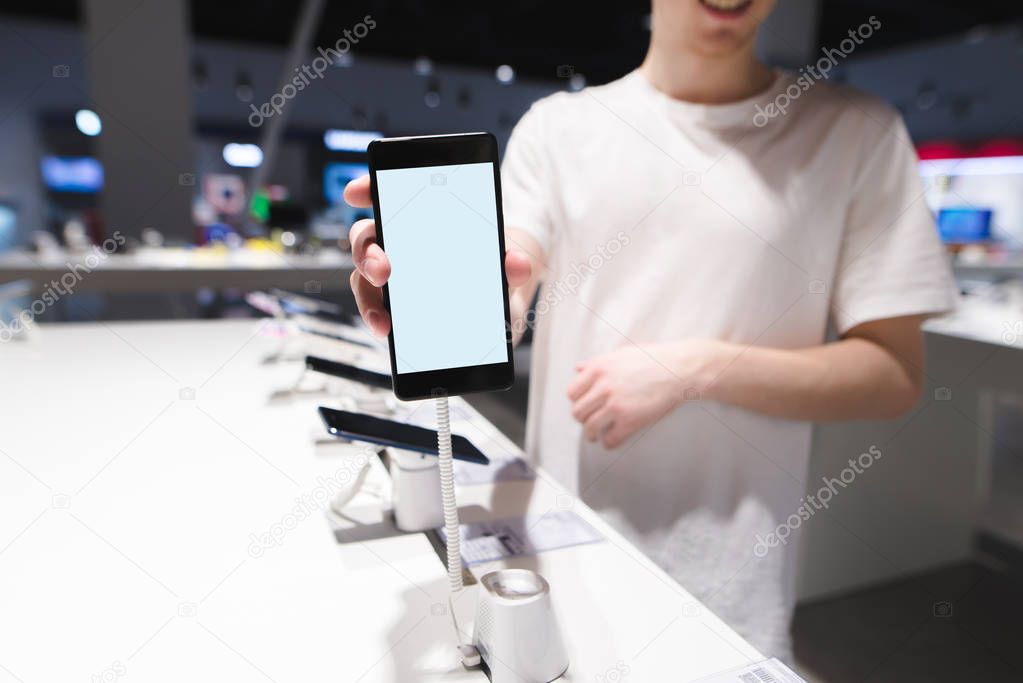 A man shows a smartphone with a white screen in the camera. Phone with a white screen on the background of the electronicsstore. Buy a phone in the store.