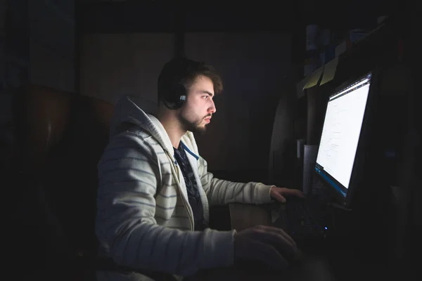 Молодой человек с бородой и наушниками ночью пользуется интернетом. Работа на домашнем компьютере ночью — стоковое фото