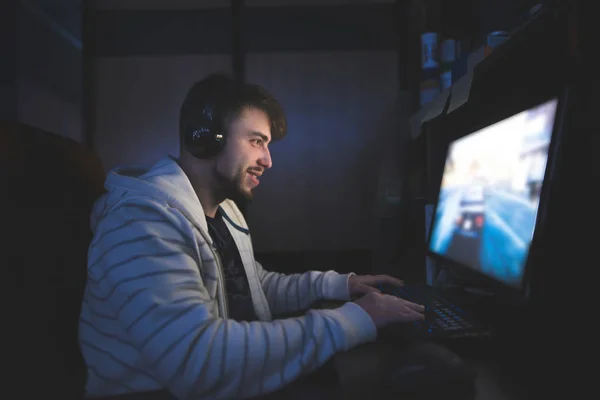 Положительный парень играет в игры за компьютером по ночам и улыбается. Молодой человек играет в компьютерные игры дома в своей комнате — стоковое фото