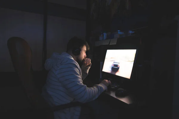 De jonge man in de koptelefoon zit op de computer en het spel speelt. De gamer maakt gebruik van de computer's nachts. Racen op de computer. Gamer concept. — Stockfoto